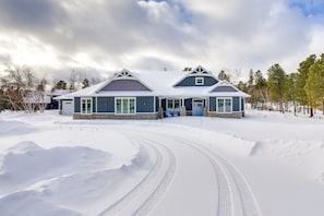 Uma casa grande de dois andares com um caminho de acesso coberto de neve e paisagem ao redor.