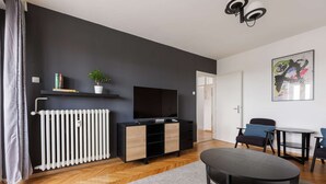 A modern living room with a black wall, white radiator, wooden floor, a black sofa, a small table, and a TV on a black stand.