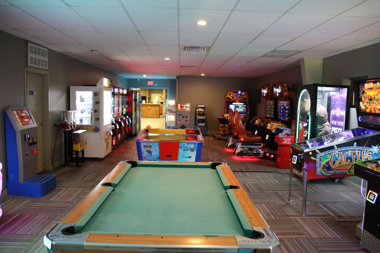 A pool table in the center of a room with arcade games and a vending machine on the left.