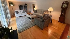A living room with a white sofa, a grey sofa, a plaid armchair, a wooden floor, and a large clock on the wall. - Hidden oasis with northern feel (Cloquet)