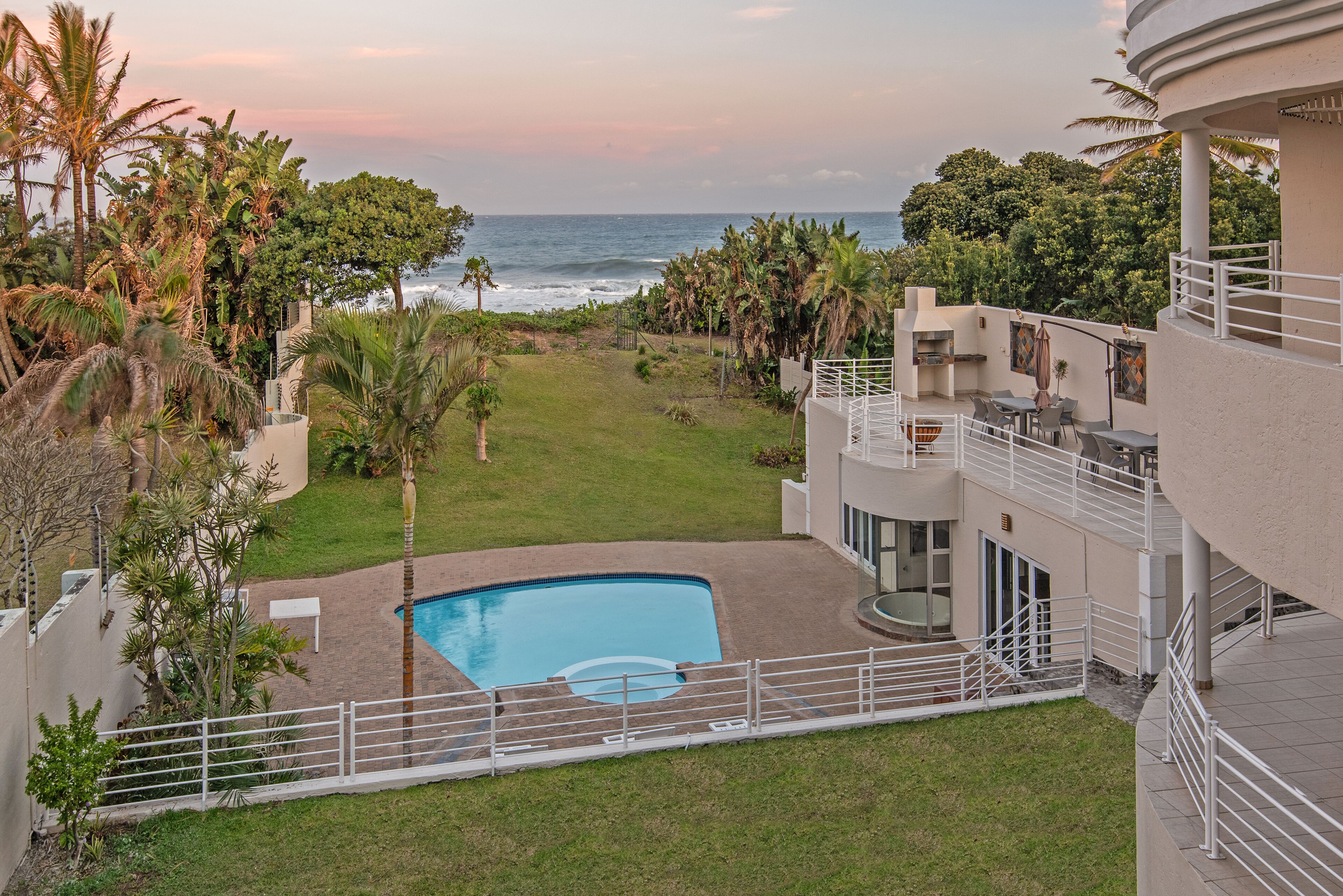 A pool area with a view of the ocean, surrounded by palm trees and a building with outdoor seating.