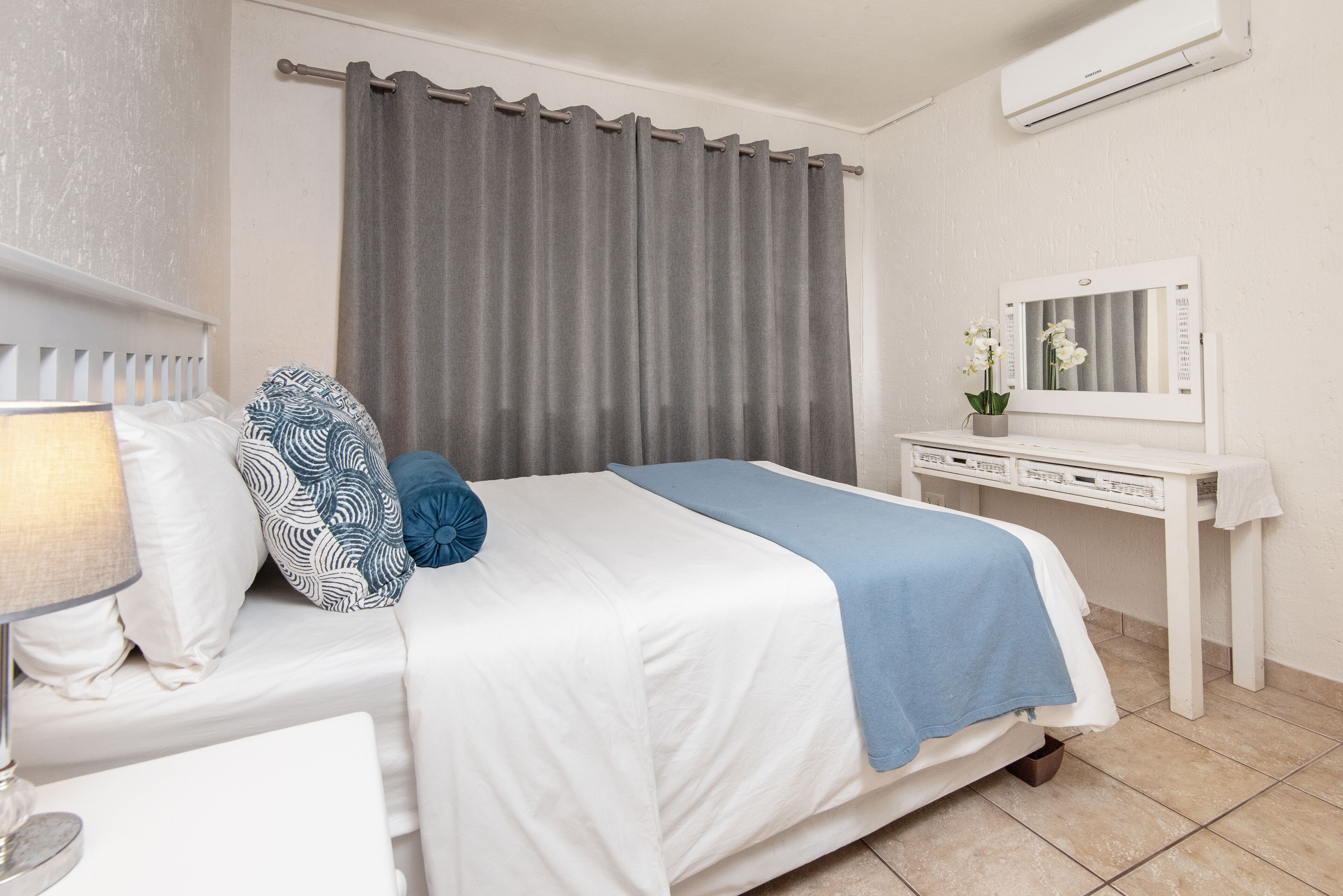 A neatly arranged bedroom with a bed, a bedside table, a lamp, a dresser, and a mirror.