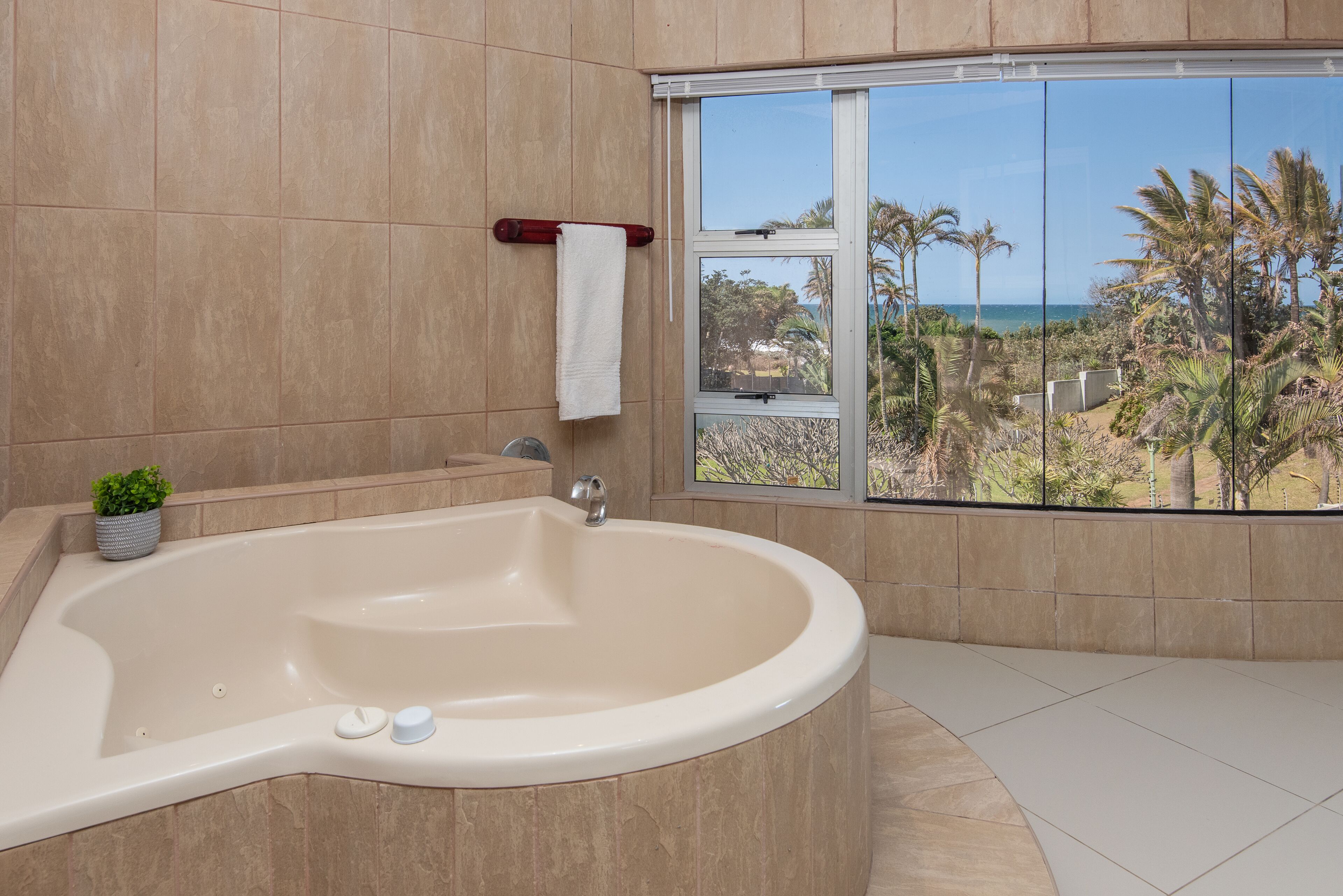 A bathroom with a large bathtub, a window with a view of palm trees and the ocean, and a towel rack with a white towel.
