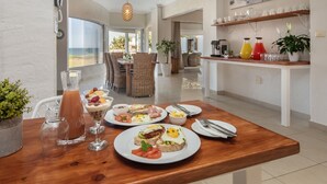 Una mesa de desayuno con diversos platos, vista a la playa y una decoración interior moderna.