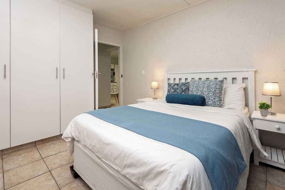 Quarto com cama, guarda-roupa branco, mesas de cabeceira com luminárias e uma porta que dá para outro cômodo.