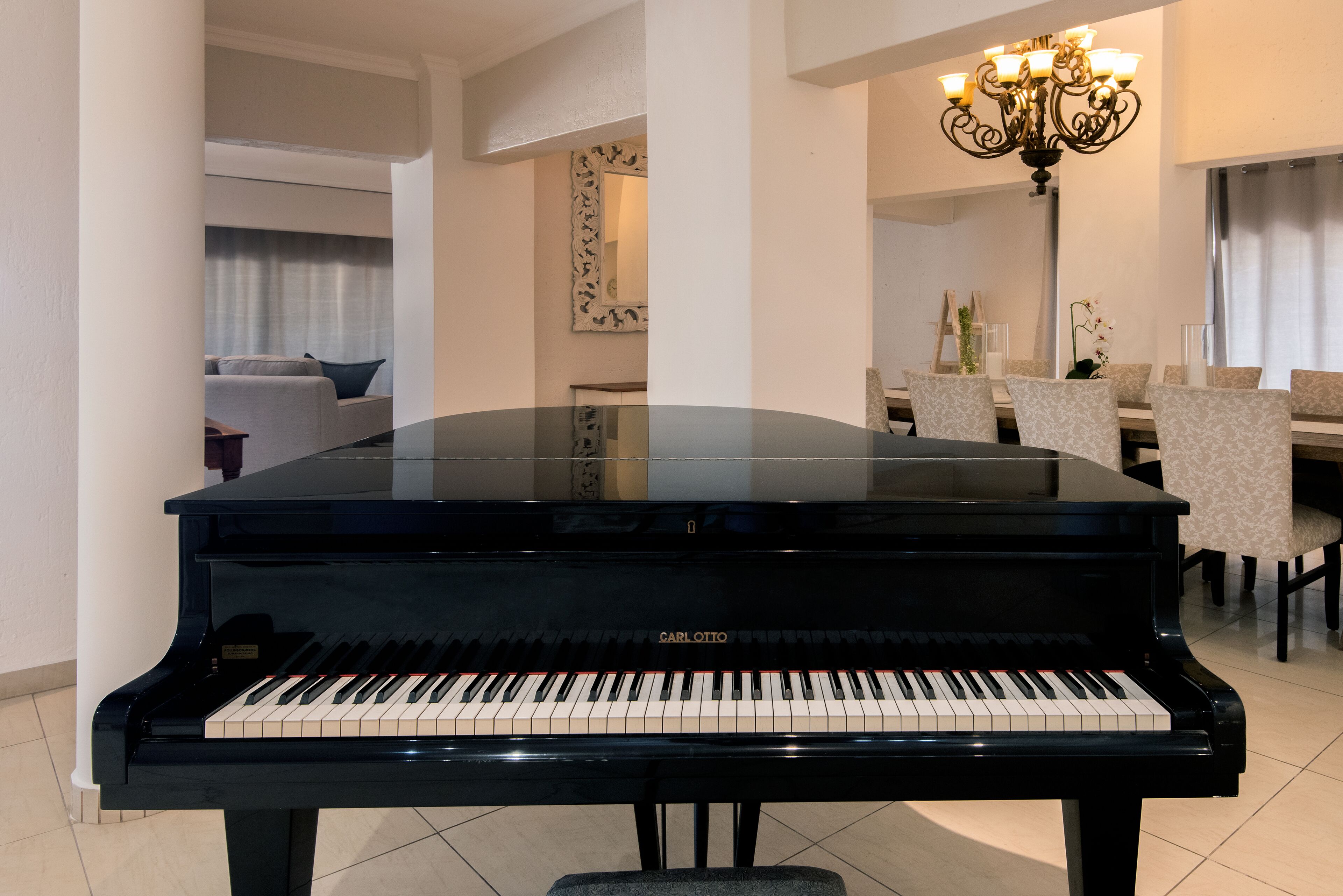 A black grand piano in a well-lit room with a chandelier, a dining area with chairs, and a sofa in the background.