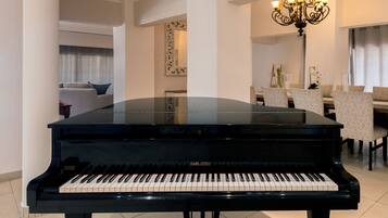 A black grand piano in a well-lit room with a chandelier, a dining area with chairs, and a sofa in the background.