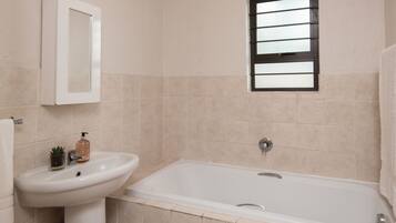 A bathroom with a sink, a mirror, a window, and a bathtub.