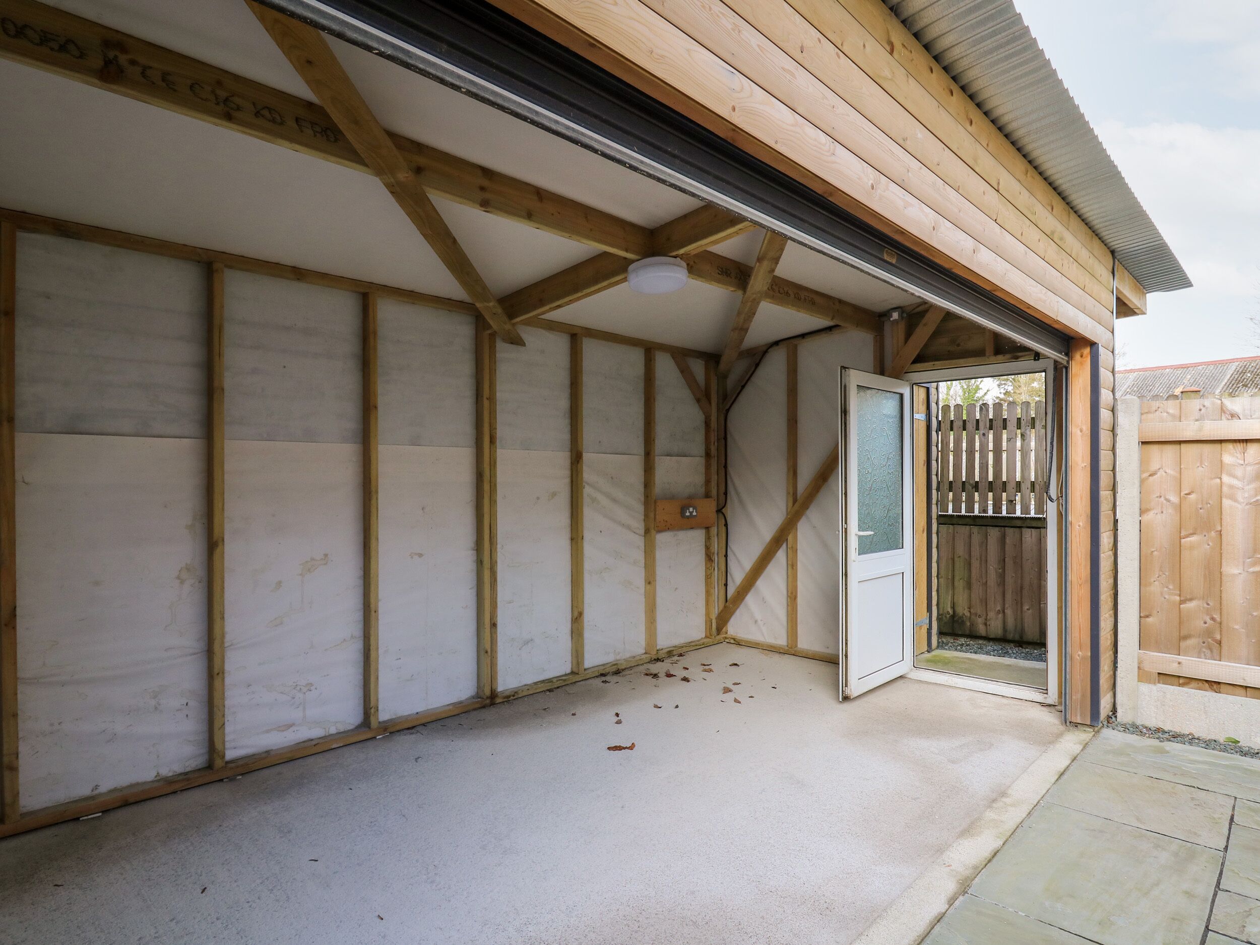 A building under construction with exposed wooden beams and a partially open door leading to a fenced area.