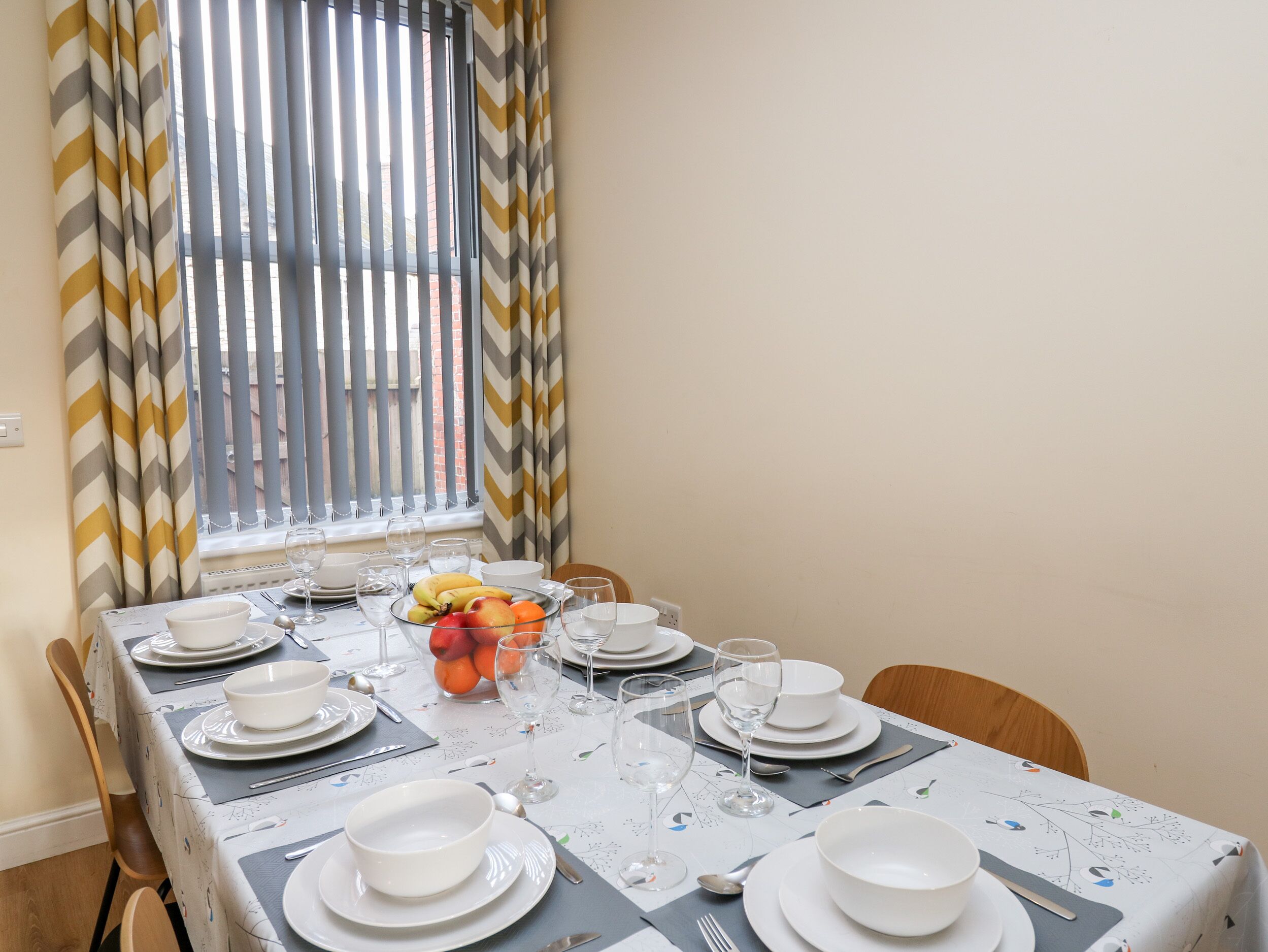 Mesa de comedor dispuesta para cuatro personas, con platos, tazones y cubiertos blancos, un frutero y copas de vino.