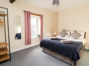 A bedroom with a bed, a bedside table with a lamp, a window with curtains, a mirror, and a shelf with a towel. - Afonwy House (Rhayader)