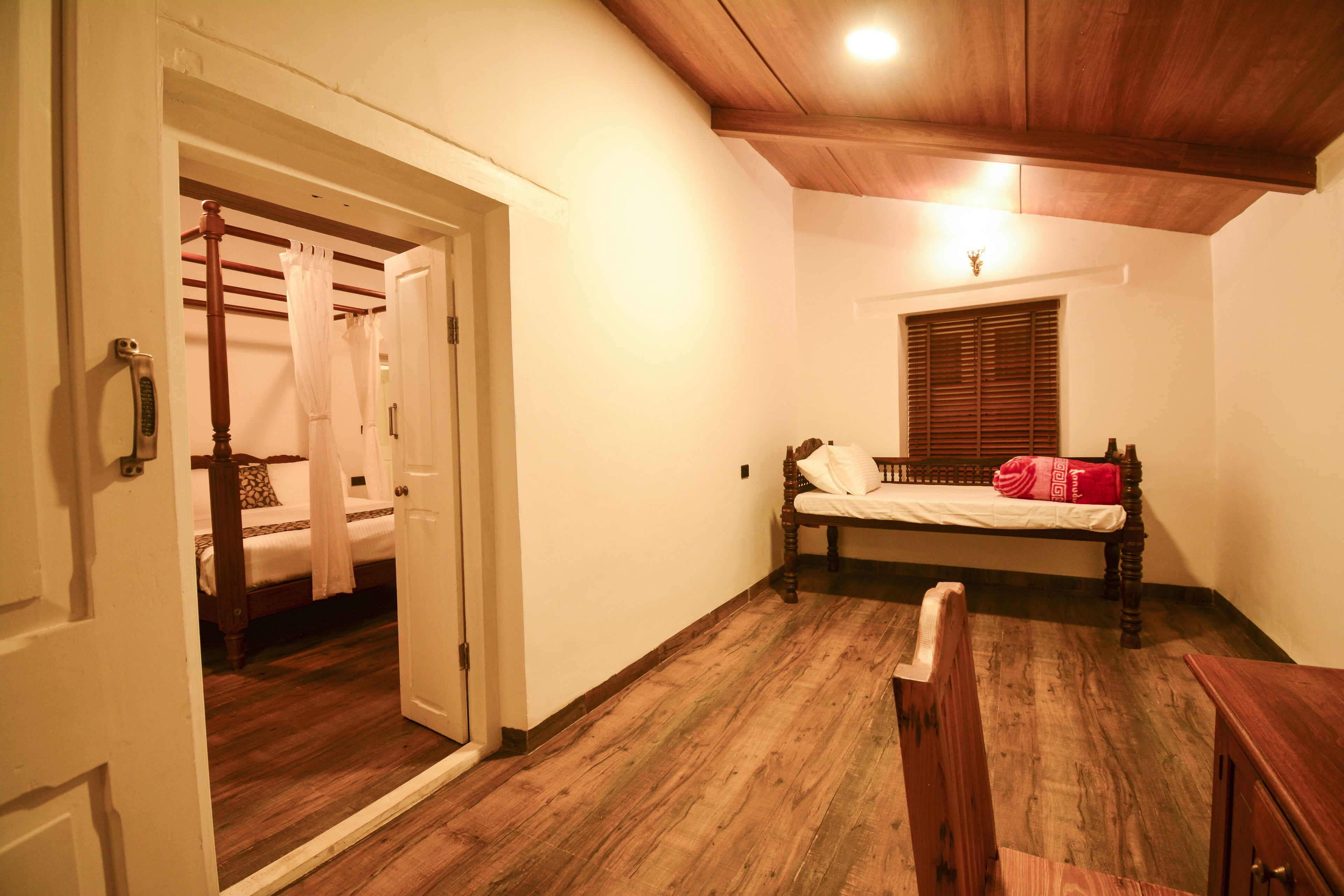 A room with a wooden floor, a bed with a white mattress and a red blanket, a wooden bench, and a window with wooden blinds.