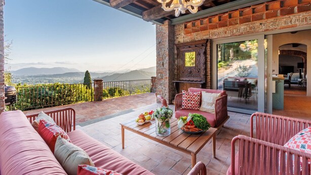 A cozy outdoor seating area with a view of distant mountains, a brick fireplace, and a modern interior. - Villa Alice, panoramic stone farmhouse to sleep 10 with pool in Lucca (Lucca)