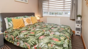 Une chambre à coucher comprenant un lit, une table de chevet, une lampe, une fenêtre avec des stores, un radiateur, une petite plante et un tableau encadré accroché au mur.