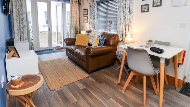 Un salon chaleureux avec un canapé en cuir marron, une table basse blanche et une table à manger avec des chaises. Il y a une grande fenêtre avec des rideaux, une porte donnant sur l’extérieur et une petite table d’appoint en bois.