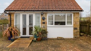 Une maison à deux étages, avec un toit recouvert de tuiles rouges, une porte de garage et une porte d’entrée dotée de panneaux de verre. Des plantes en pot sont disposées devant la maison, et un petit panneau est posé sur le sol.