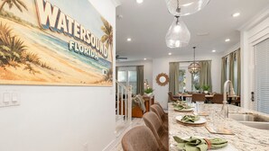 A dining area with a long table, chairs, and a kitchen area with a sink and countertop. - 🌅 Sunset Palms | Golf Cart, Retro Game Room & Walk to The Big Chill (Inlet Bech)