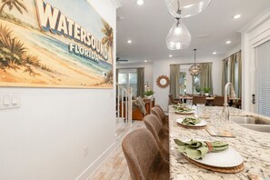 A dining area with a long table, chairs, and a kitchen area with a sink and countertop. - 🌅 Sunset Palms | Golf Cart, Retro Game Room & Walk to The Big Chill (Inlet Bech)