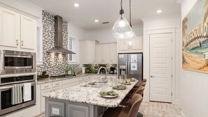 A modern kitchen with white cabinetry, a granite island, stainless steel appliances, and a hanging light fixture. - 🌅 Sunset Palms | Golf Cart, Retro Game Room & Walk to The Big Chill (Inlet Bech)