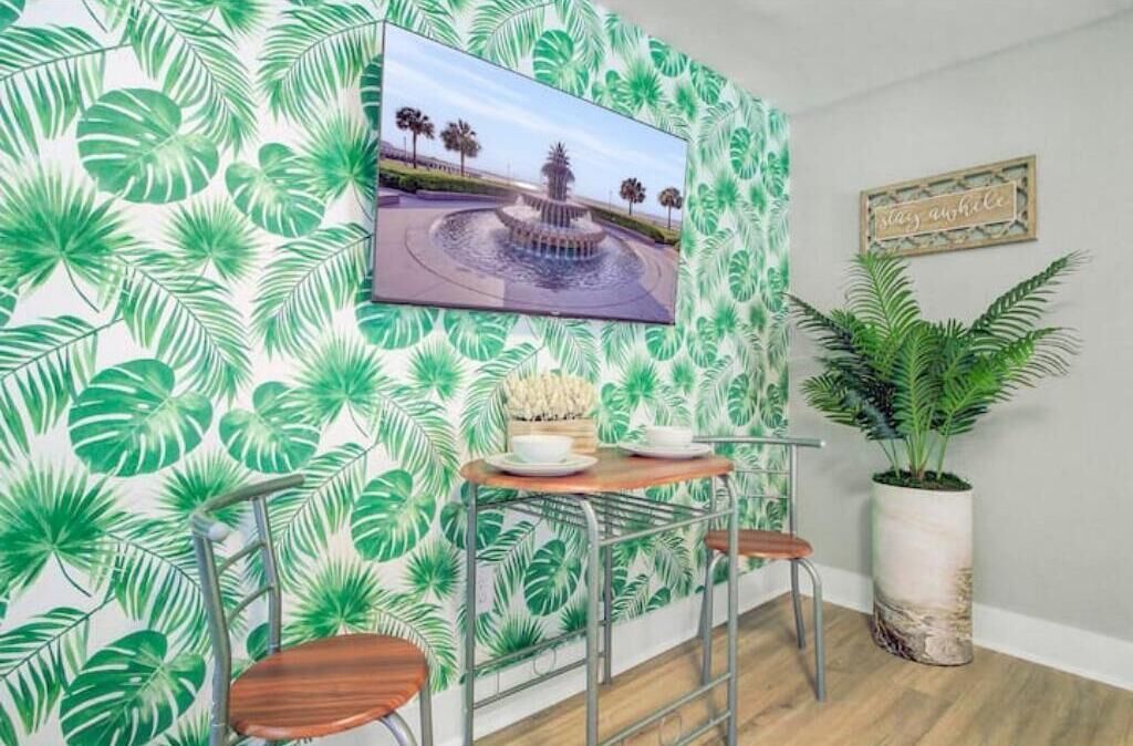 Una stanza con carta da parati a tema tropicale, una TV a parete, un piccolo tavolo di legno con sedie e una pianta in vaso.