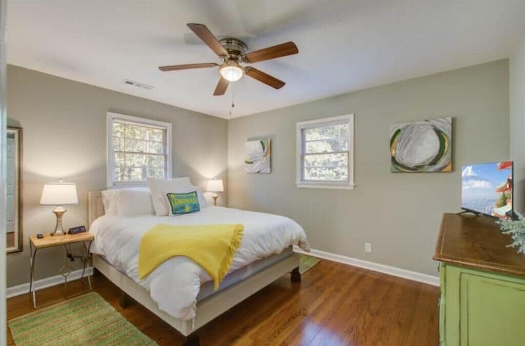 A bedroom with a bed, a ceiling fan, two windows, a nightstand, a lamp, and a television on a cabinet.
