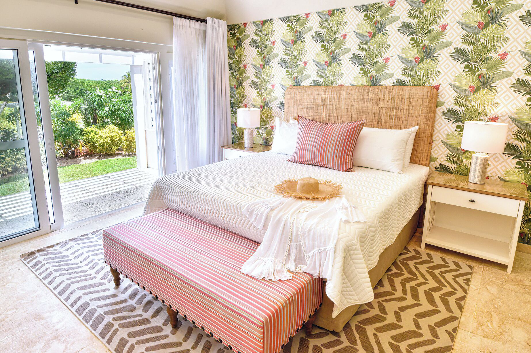A bedroom with a large bed, a bench, a nightstand, and a view of greenery through a sliding door.