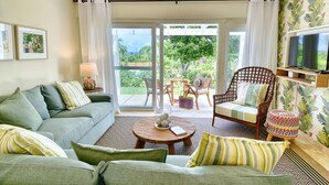 Ein Wohnzimmer mit einer Couch, Sesseln, einem Couchtisch und einem Fernseher. Eine Glastür öffnet sich zu einem Außenbereich mit einem Sessel und einem Tisch.