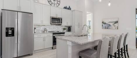 A modern kitchen with white cabinetry, stainless steel appliances, a central island with bar stools, and a clock on the wall.