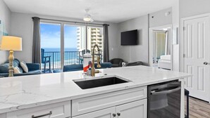 A modern kitchen with a white marble countertop, a built-in dishwasher, and a view of the ocean through large windows. - 9th-Floor Gulf Sunset Views | 2 King Beds | Shores of Panama 920 (Panama City Beach)