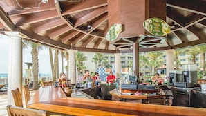 An outdoor bar area with wooden tables and chairs, a counter with a cash register, and a view of the beach and palm trees. - 9th-Floor Gulf Sunset Views | 2 King Beds | Shores of Panama 920 (Panama City Beach)
