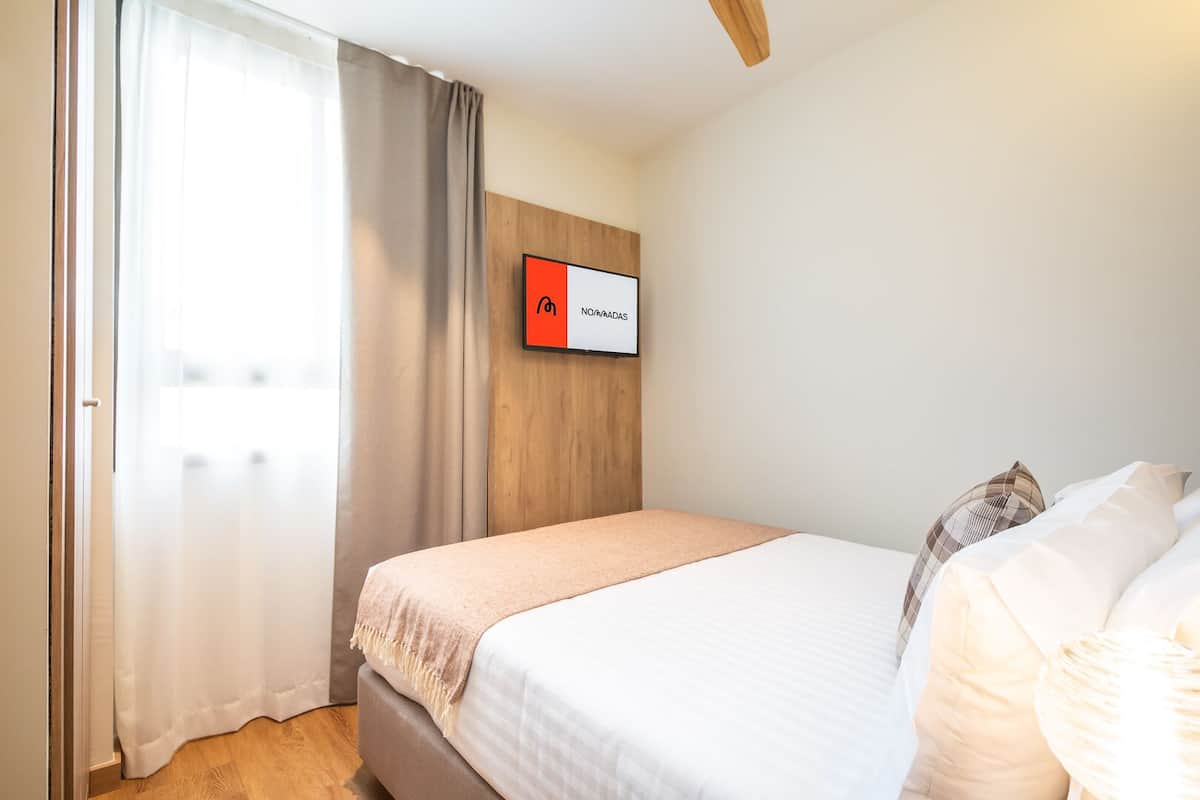 A hotel room with a bed, a TV mounted on the wall, a window with curtains, and a wooden ceiling.