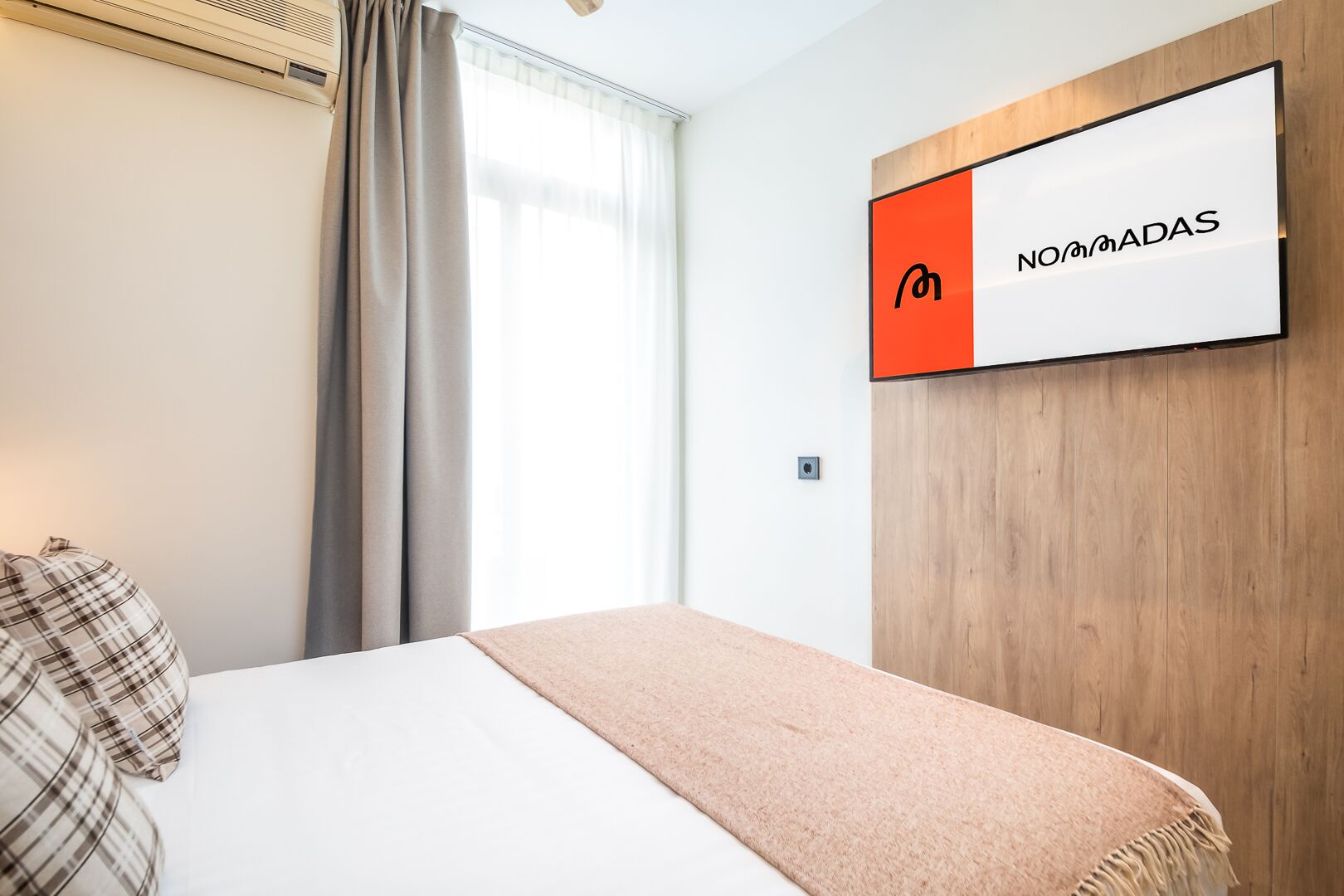 Une chambre d’hôtel avec un lit, un coussin à carreaux, un plaid beige, une fenêtre avec des rideaux et une enseigne fixée au mur portant l’inscription « NOMADAS ».
