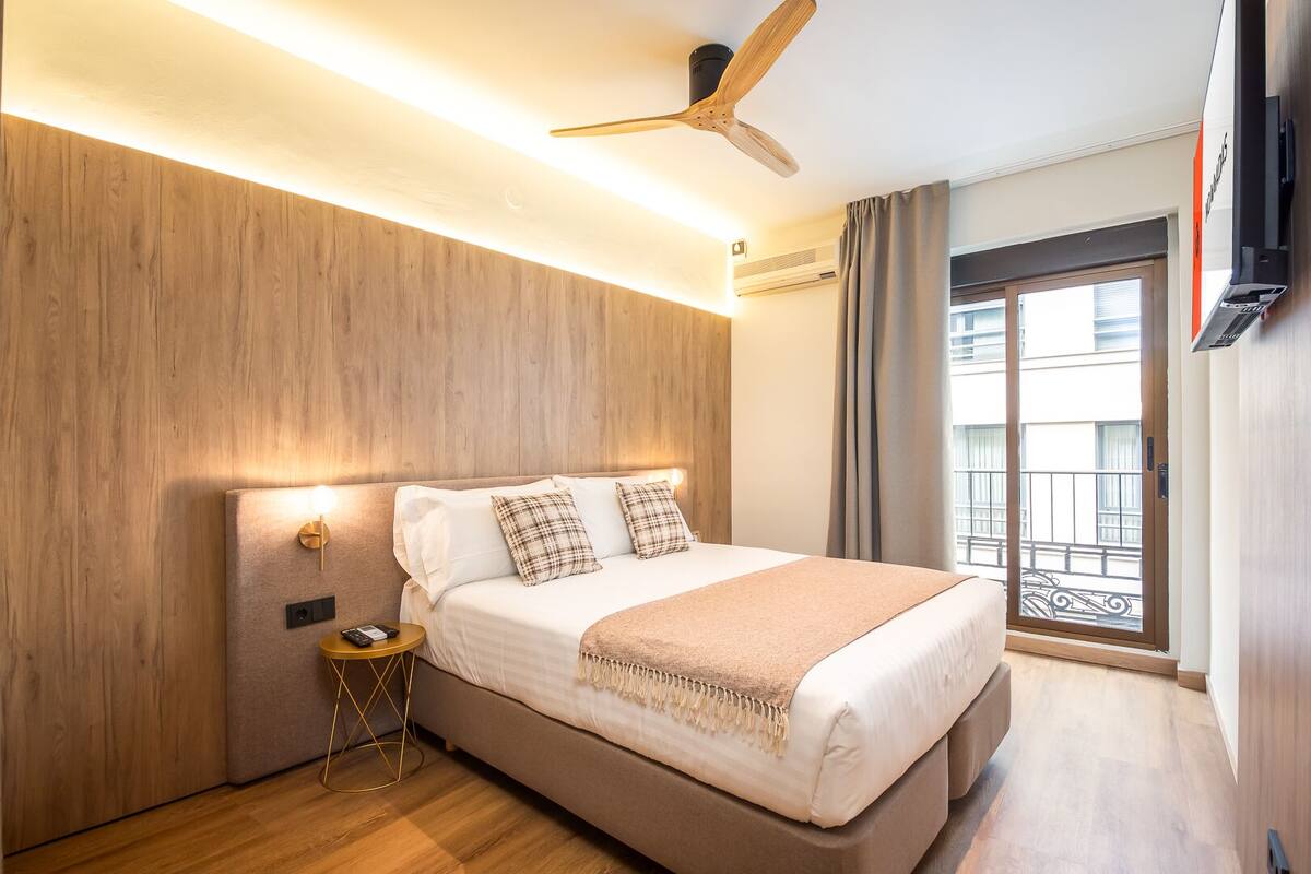 A modern hotel room with a wooden headboard, a ceiling fan, a bedside table with a lamp, and a large window with a view of the city.