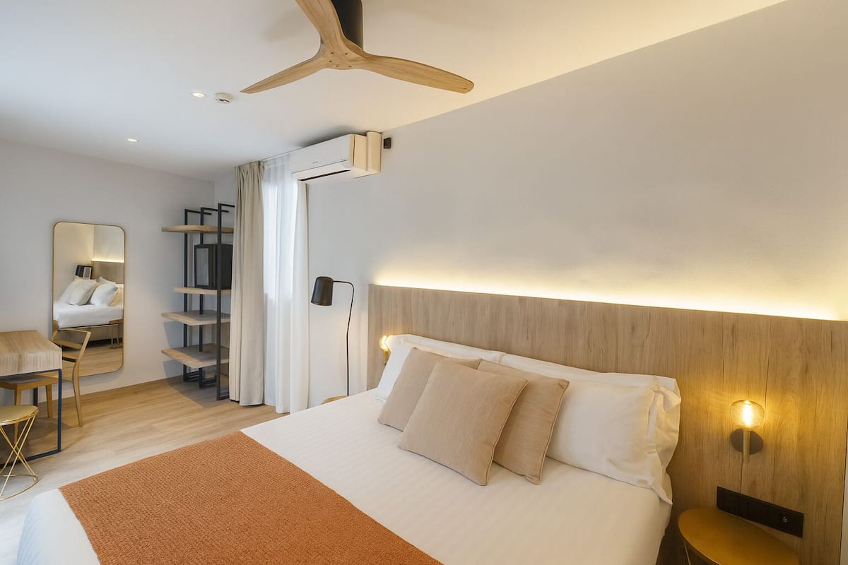 A modern hotel room with a large bed, a wooden headboard, a ceiling fan, a bedside table, and a mirror.