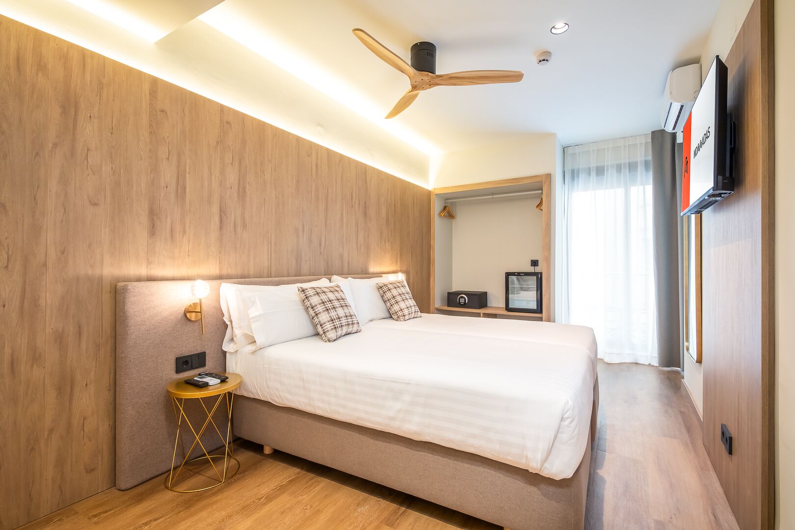 A modern hotel room with a wooden accent wall, a bed with white bedding, a round gold side table, and a ceiling fan.