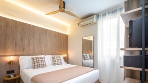A modern bedroom with a wooden headboard, a bed with white linens and plaid pillows, a bedside table with a lamp, a ceiling fan, an air conditioning unit, and a TV on a wooden shelf. - Nommadas Jero Hotel - Adults Only (Valencia)