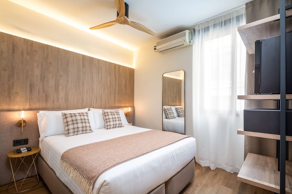 A modern bedroom with a wooden headboard, a bed with white linens and plaid pillows, a bedside table with a lamp, a ceiling fan, an air conditioning unit, and a TV on a wooden shelf.