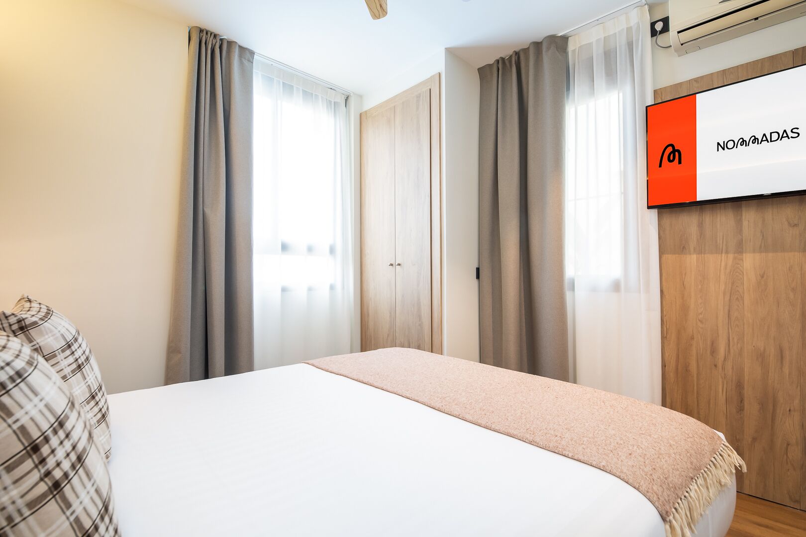 A hotel room with a bed, curtains, a wooden wardrobe, and a wall-mounted TV displaying "NO MADAS".