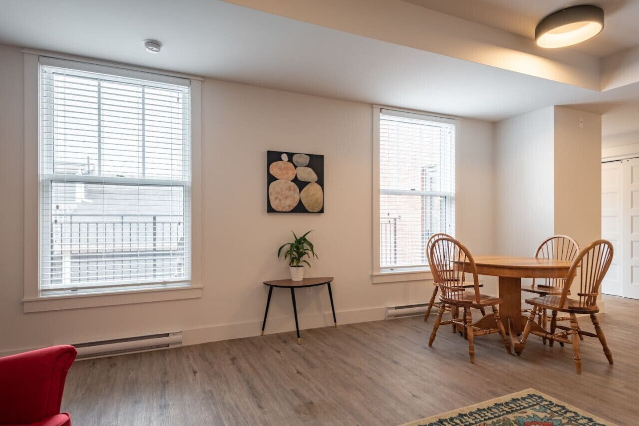 Un coin repas avec une table et des chaises en bois, de grandes fenêtres équipées de stores, une petite plante en pot et une œuvre d’art accrochée au mur.