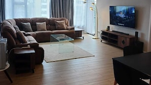 Ett modernt vardagsrum med en brun lädersoffa, ett soffbord i glas, ett trägolv, en platt-TV och en matplats med ett svart bord och stolar.