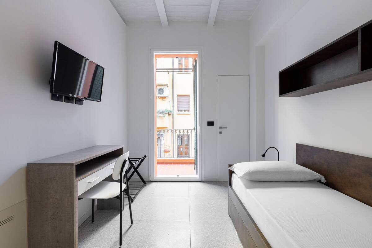 Une petite chambre moderne, équipée d’un lit, d’un bureau et d’un téléviseur fixé au mur.