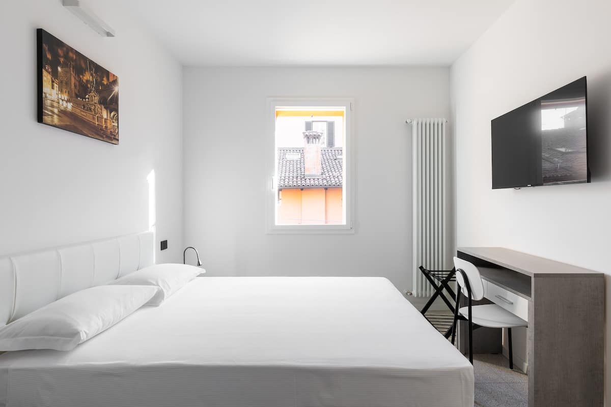 Une chambre d’hôtel équipée d’un lit, d’un bureau, d’une chaise, d’une télévision et d’une fenêtre donnant sur des immeubles.