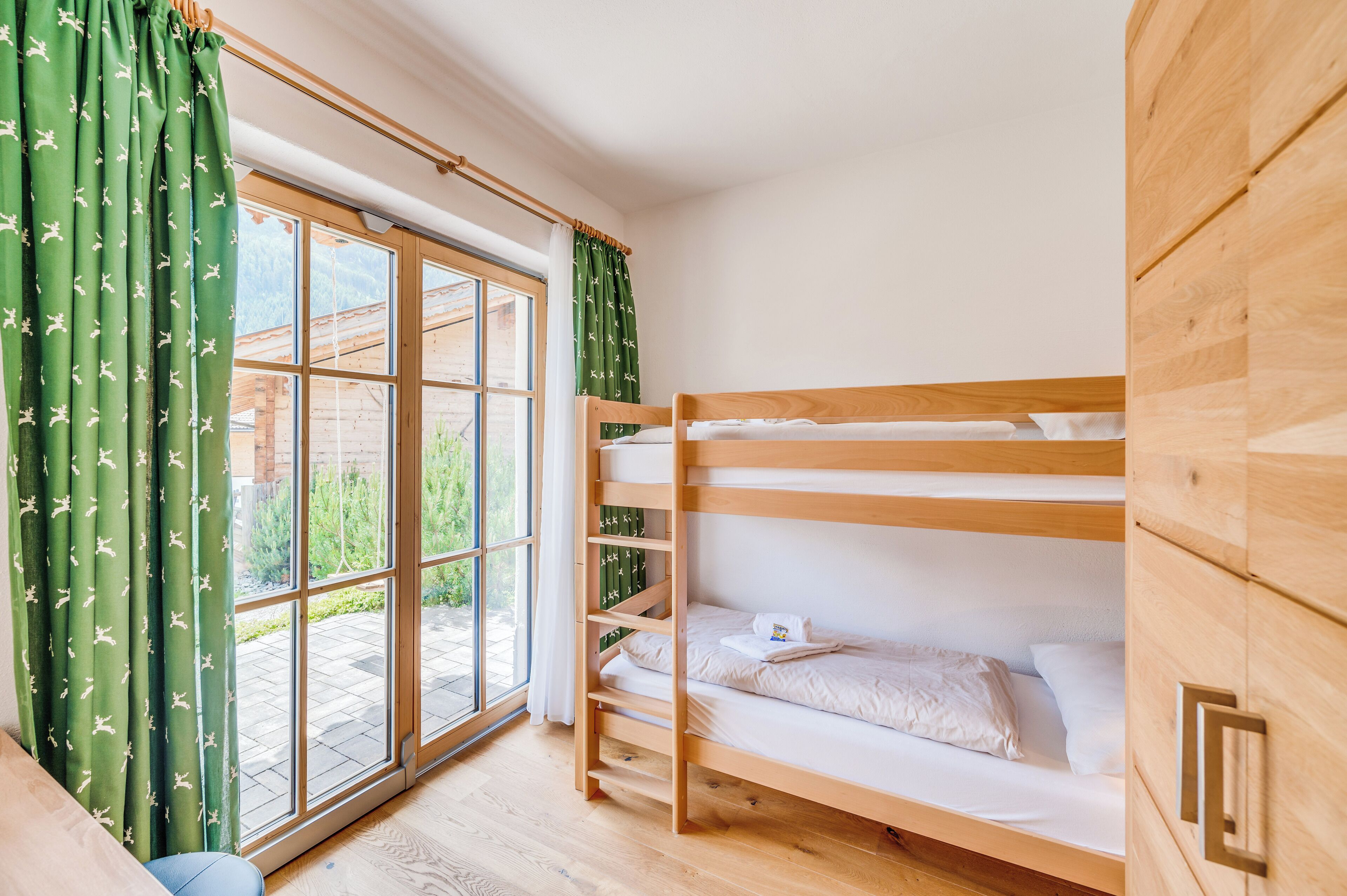 Quarto com beliche, piso de madeira, janela com cortinas e guarda-roupa de madeira.