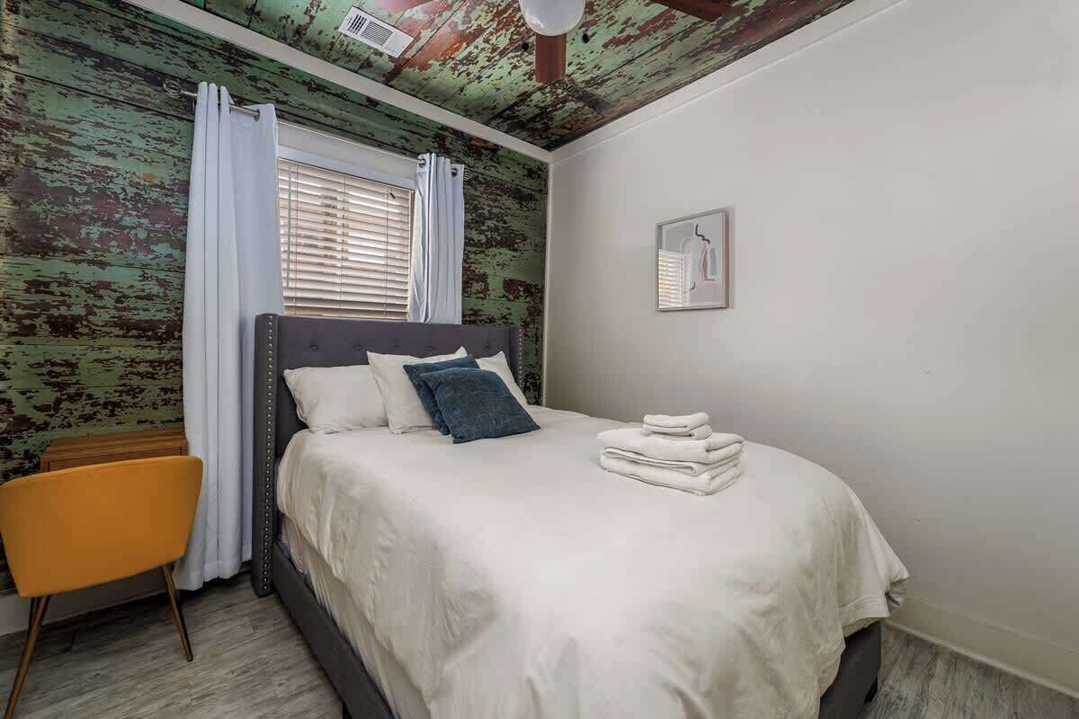 A bedroom with a bed, a chair, a window with curtains, and a wall with a distressed finish.