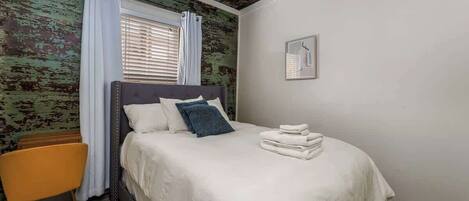 A bedroom with a bed, a chair, a window with curtains, and a wall with a distressed finish.