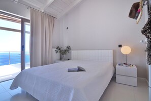 Un dormitorio con una cama grande, una mesita de noche con lámpara y vista al mar a través de una puerta corredera.