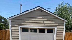 A single-story shed with a white garage door, surrounded by a wooden fence, and a concrete driveway leading to it.