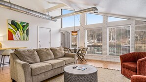 A spacious living room with a grey sofa, a round coffee table, and a red armchair. There is a dining area with a table and chairs, and large windows providing natural light. - Riverfront apartment downtown with parking (Saugatuck)