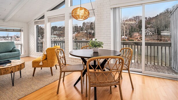 A modern living room with a round dining table, wicker chairs, a blue sofa, and large windows overlooking a body of water. - Downtown apartment with water view and parking (Saugatuck)