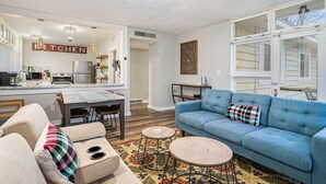 Una sala moderna con un sofá azul, un sofá color beige y una mesa de centro. Se observa una cocina al fondo con un letrero que dice "KITCHEN".